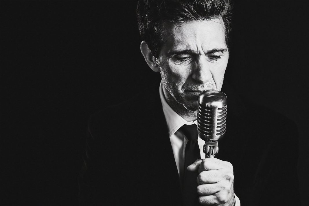 black and white photo of Ronnie Marmo in a suit with his hand on a a vintage stand microphone.