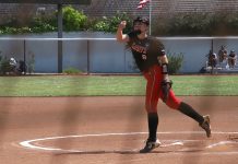 Saint Martin's University softball player in a red and black uniform. She in side a white-chalk outlined circle on red dirt with her arm outstretch and a leg back, as fi she had just thrown a ball. her other hand has a catcher's mitt on